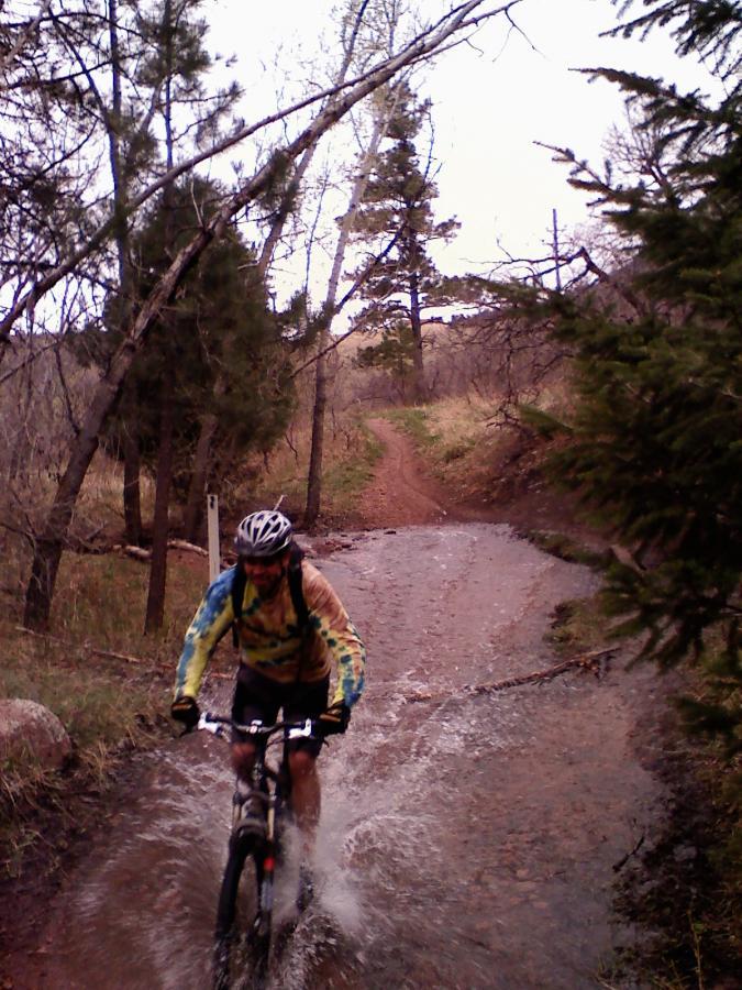 Mountain biker riding through a puddle on a dirt trail surrounded by trees, creating a splash of water. The trail is flanked by greenery, and the weather appears overcast. Falcon Trail mountain bike trail.