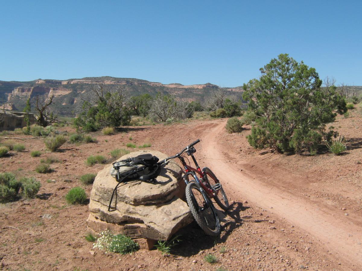 Mountain bike resting on a rock beside a dirt trail, surrounded by sparse vegetation and distant mountains under a clear blue sky. Mary's Loop / Horsethief Bench mountain bike trail.