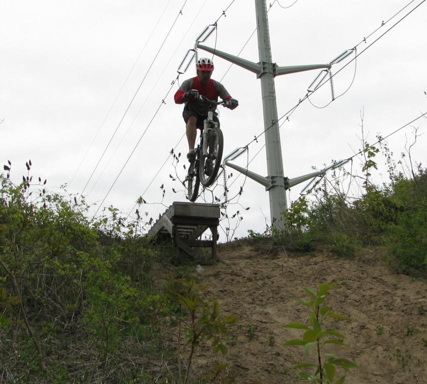 A person in a red helmet and shirt is jumping off a wooden ramp while riding a mountain bike, with power lines visible in the background and a hilly, grassy terrain beneath. The sky is overcast, suggesting a cloudy day. Don Valley mountain bike trail.