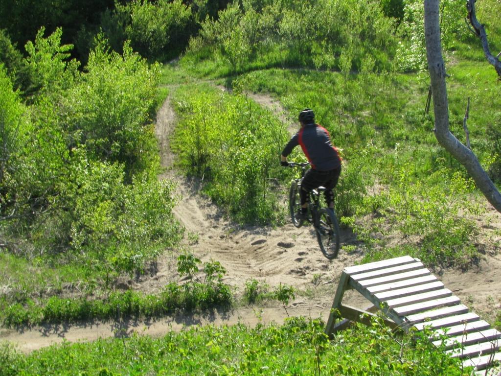 A mountain biker in a black helmet and gray-red clothing jumps off a wooden ramp on a dirt trail surrounded by lush green vegetation and small trees. The scene captures the thrill of mountain biking in a natural outdoor setting. Don Valley mountain bike trail.