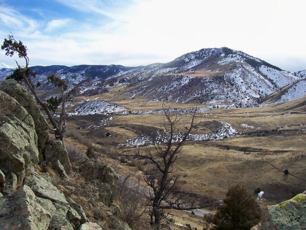 A panoramic view of rolling hills and mountains, featuring a mix of rocky terrain and patches of snow. In the foreground, a rocky outcrop and a gnarled tree branch extend into the scene. The landscape includes open fields with dried grass and winding paths, under a cloudy sky with hints of blue peeking through. Red Rocks / Dakota Ridge mountain bike trail.