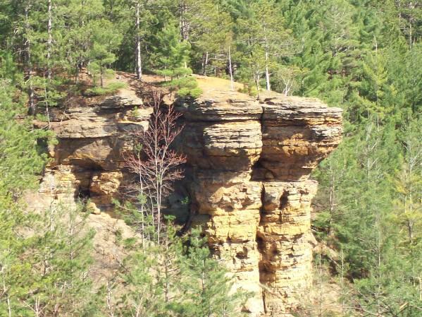 A large, rocky outcrop surrounded by lush pine trees, featuring distinct layers of stone that form a prominent cliff-like structure. Levis Mounds mountain bike trail.