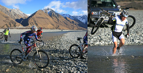 A scenic outdoor setting featuring several mountain bikers crossing a rocky riverbed. On the left, cyclists navigate through shallow water, surrounded by stunning mountains and a clear blue sky. On the right, a biker is lifting their bike over their shoulder while walking through the water.