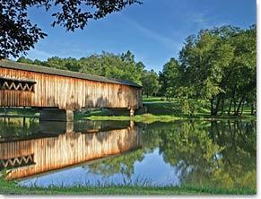 A picturesque scene featuring a wooden covered bridge spanning a calm river, surrounded by lush greenery and trees under a clear blue sky. The water reflects the bridge and surrounding landscape, creating a serene and tranquil atmosphere. Watson Mill Bridge State Park mountain bike trail.