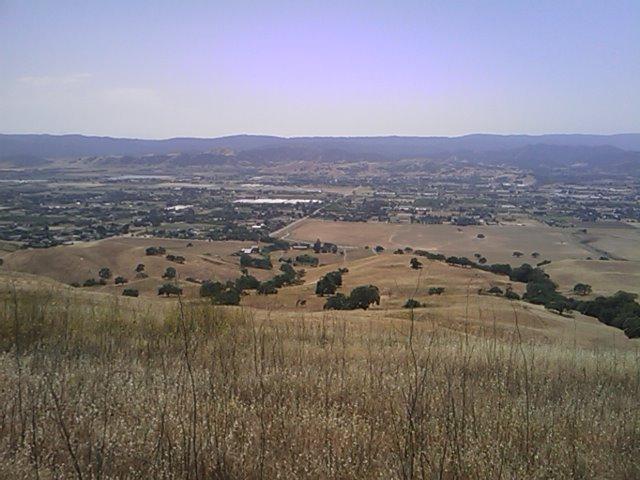 A panoramic view of rolling hills and valleys under a clear blue sky, showcasing a mix of dry grassland and patches of green vegetation. In the distance, a sprawling valley is dotted with trees and structures, separated by gentle slopes. The scene captures the natural beauty of a rural landscape. Harvey Bear Trail mountain bike trail.