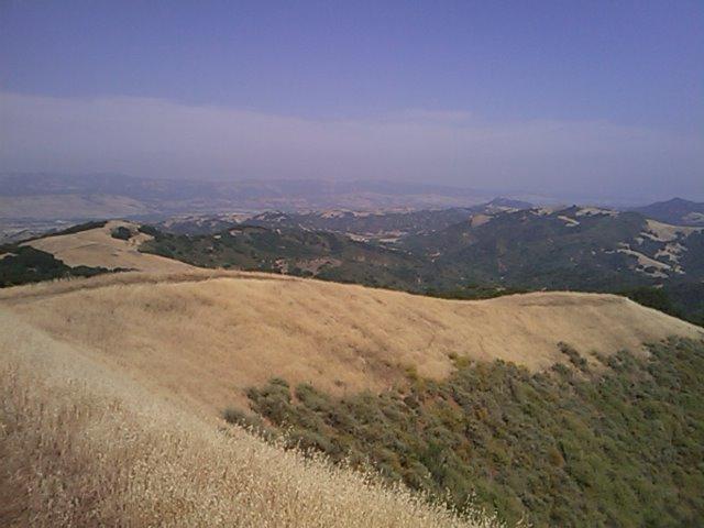 A panoramic view of rolling hills covered with dry grass and patches of greenery, under a clear blue sky with distant mountains in the background. The landscape showcases a mix of yellow and green hues typical of a warm season. Rancho Canada Del Oro mountain bike trail.