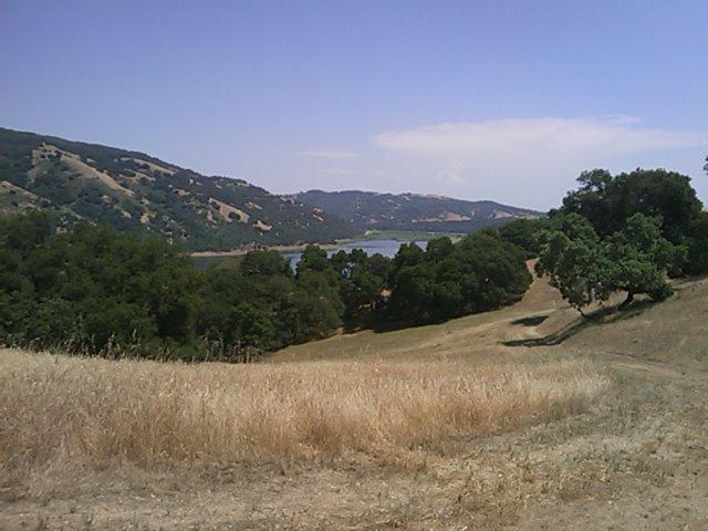 A scenic view of rolling hills and a tranquil lake, surrounded by lush greenery and dry grass. The landscape features a mix of trees and open fields, with distant mountains under a clear blue sky. Harvey Bear Trail mountain bike trail.