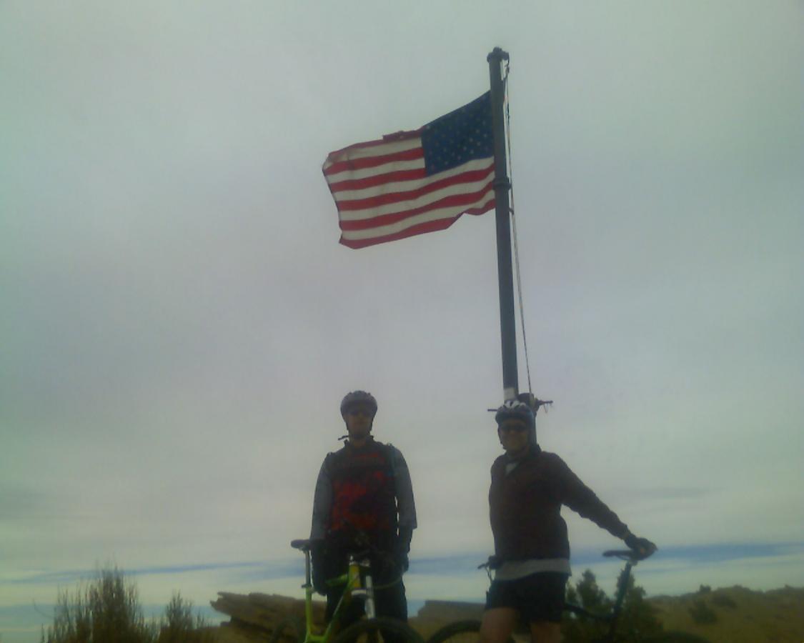 Two mountain bikers pose with their bicycles near a tall flagpole displaying the American flag against a cloudy sky. The scene captures a sense of adventure and patriotism. Stagecoach mountain bike trail.