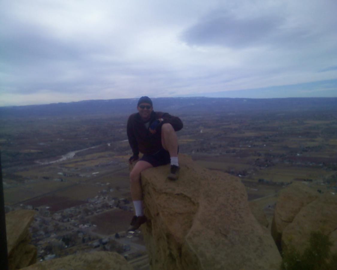 A person sitting on a large rock outcrop at a mountain summit, dressed in casual outdoor clothing, looking out over a scenic landscape with valleys and mountains in the background under a cloudy sky. Stagecoach mountain bike trail.