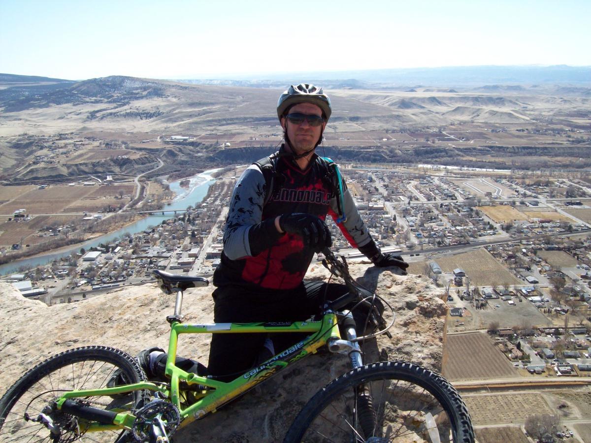 A mountain biker sitting on a rocky outcrop, with a green bicycle beside him. The expansive landscape below features a winding river, patches of farmland, and a small town against a backdrop of hills and clear blue sky. The biker is wearing protective gear, sunglasses, and a jersey, looking out over the view. Stagecoach mountain bike trail.
