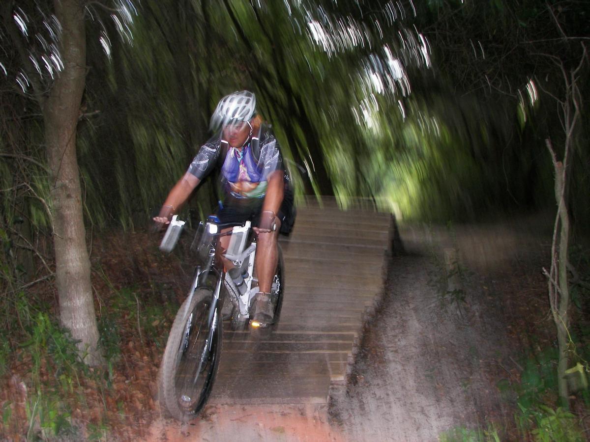 A mountain biker navigating a wooden ramp through a wooded trail, captured in motion with a blurred background, emphasizing speed and action. Loyce E. Harpe Park mountain bike trail.