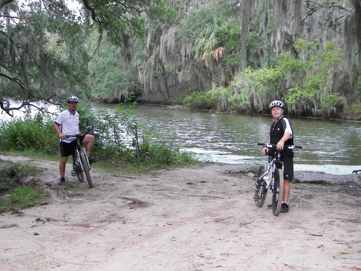 Two cyclists are standing by the edge of a river, surrounded by lush greenery and hanging Spanish moss. One cyclist, wearing a white shirt and black shorts, is resting on his bike with one leg raised. The other cyclist, dressed in a black and white outfit, stands beside his bike, both smiling at the camera. The sandy path next to the water suggests a recent ride through the natural landscape. Loyce E. Harpe Park mountain bike trail.