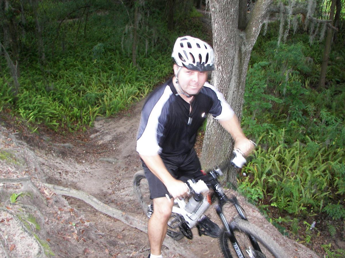 A mountain biker navigating a dirt trail surrounded by greenery. The rider, wearing a black and white cycling outfit and a helmet, is in motion, maneuvering over a rugged path with roots and loose soil. Loyce E. Harpe Park mountain bike trail.