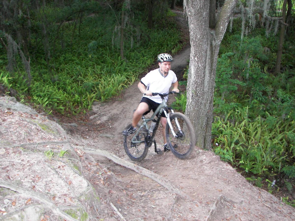 A mountain biker navigating a dirt trail in a wooded area, with greenery and trees surrounding the path. The biker is in a white shirt and black shorts, balancing on the bike as they ride downhill. Loyce E. Harpe Park mountain bike trail.