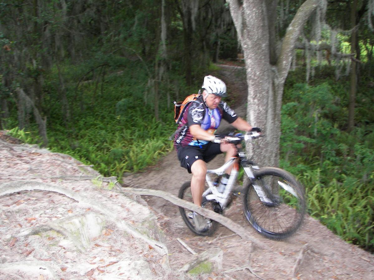 A mountain biker navigating a rugged trail, surrounded by lush greenery and trees. The cyclist is captured mid-ride, wearing a helmet and a colorful jersey, with a focus on maintaining balance on the uneven terrain. Roots and dirt are visible in the foreground, showcasing the natural setting of the biking path. Loyce E. Harpe Park mountain bike trail.
