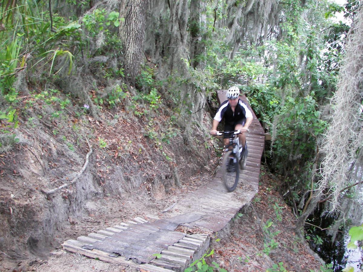 A mountain biker rides along a narrow wooden path through a lush, green forest, with trees and moss hanging nearby. The trail winds close to a body of water, showcasing a mix of natural vegetation and the rugged terrain of the area. Loyce E. Harpe Park mountain bike trail.