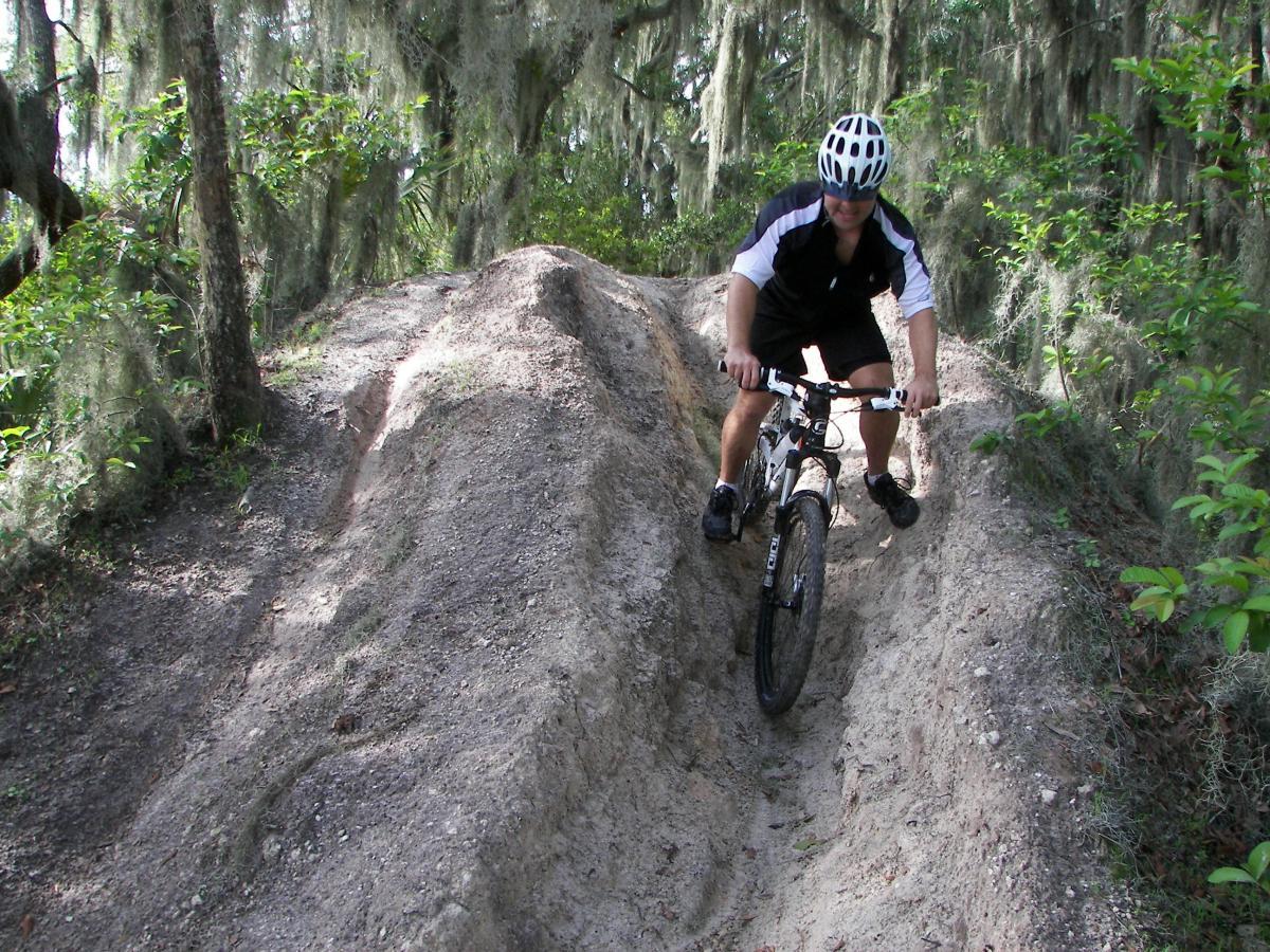 A mountain biker navigates a dirt trail in a wooded area, with moss-covered trees in the background. The biker is wearing a helmet and sports gear as they ride down a sloped path created by tire tracks. Loyce E. Harpe Park mountain bike trail.