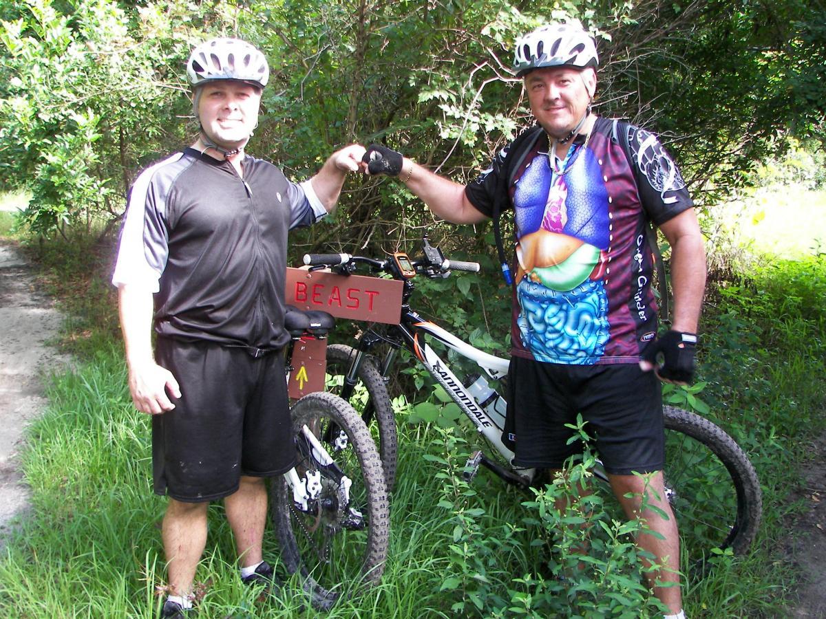 Two men in biking gear are standing beside their mountain bikes on a grassy trail. One man is wearing a black jersey and helmet, while the other is in a colorful shirt depicting an anatomical design. They are smiling and giving each other a fist bump in front of a sign that says “BEAST.” Lush greenery surrounds the area, indicating a nature trail. Loyce E. Harpe Park mountain bike trail.