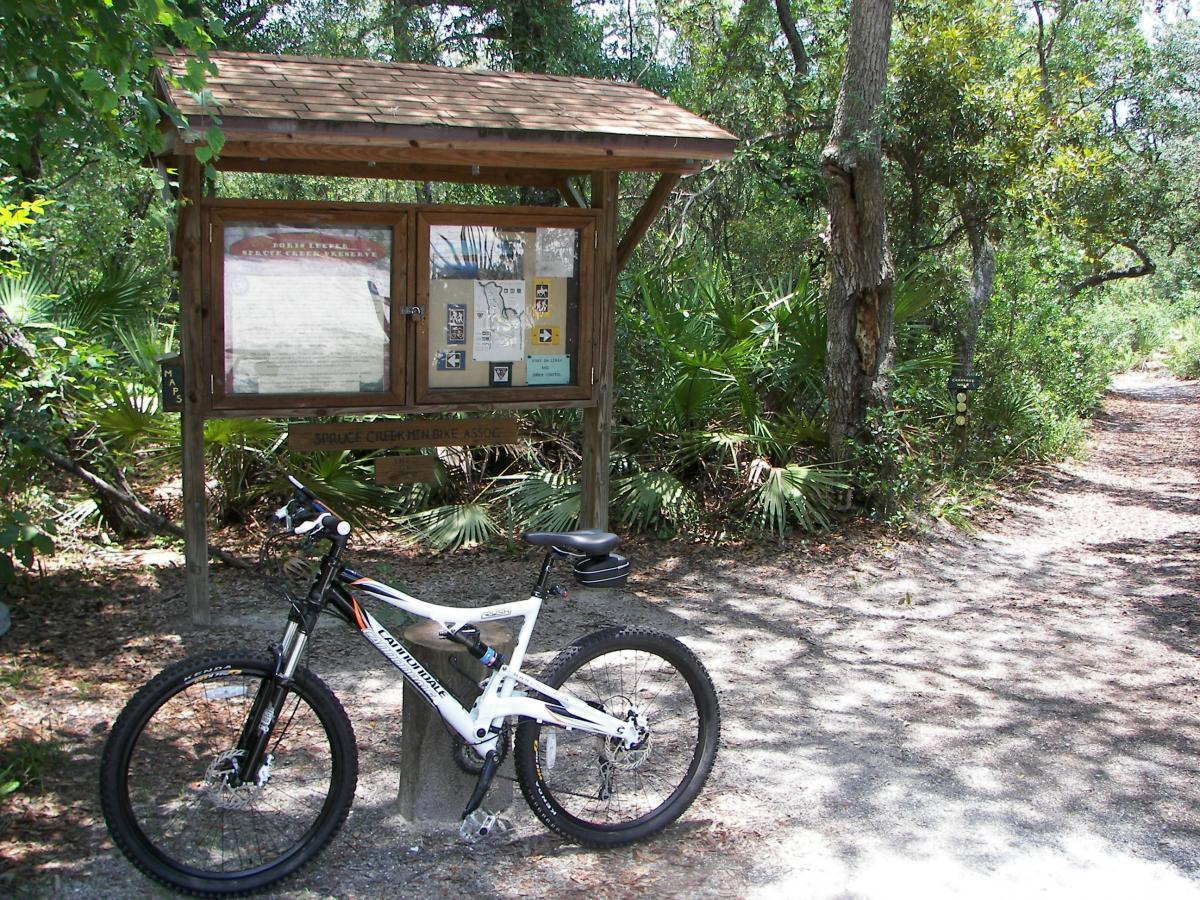 A mountain bike parked next to a wooden information kiosk surrounded by lush vegetation and trees. The kiosk features maps and trail information for Spruce Creek Mountain Bike Association, with a pathway leading into the forest on the right. Sunlight filters through the tree leaves, creating dappled shadows on the ground. Spruce Creek Preserve mountain bike trail.