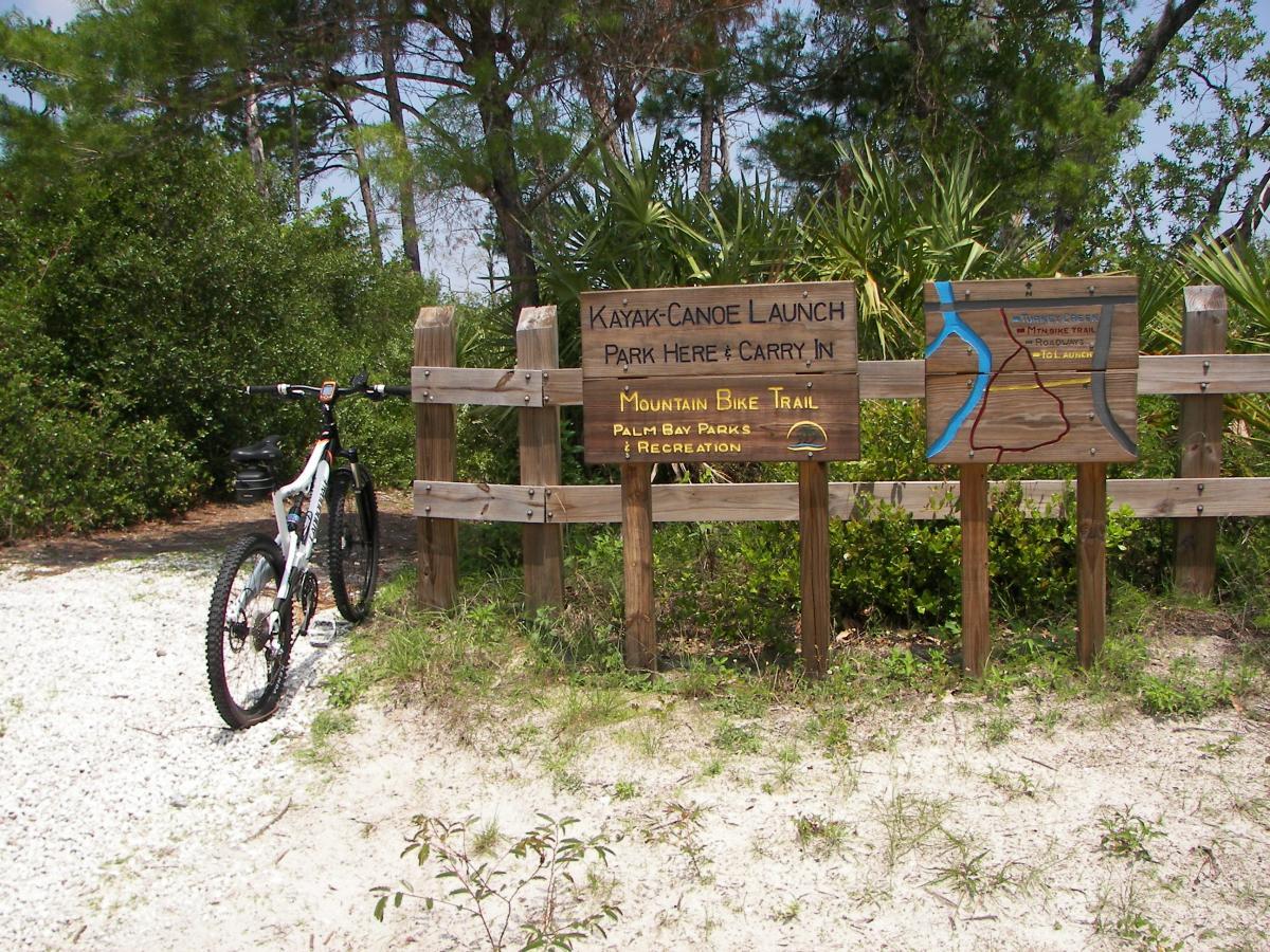 A mountain bike resting on gravel near a wooden sign indicating a kayak and canoe launch area, featuring directions to park and carry in, as well as a map of the Mountain Bay Trail at Palmetto Bay Parks and Recreation. Surrounding vegetation includes trees and shrubs. Turkey Creek mountain bike trail.