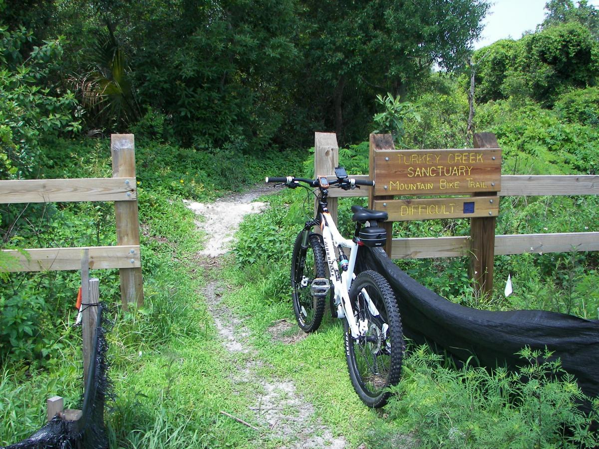 A mountain bike resting against a wooden fence at the entrance to the Turkey Creek Sanctuary Mountain Bike Trail, which is marked as difficult. The path ahead is flanked by lush greenery, with a clear division in the trail visible. A sign indicating the trail name and difficulty level is prominently displayed. Turkey Creek mountain bike trail.