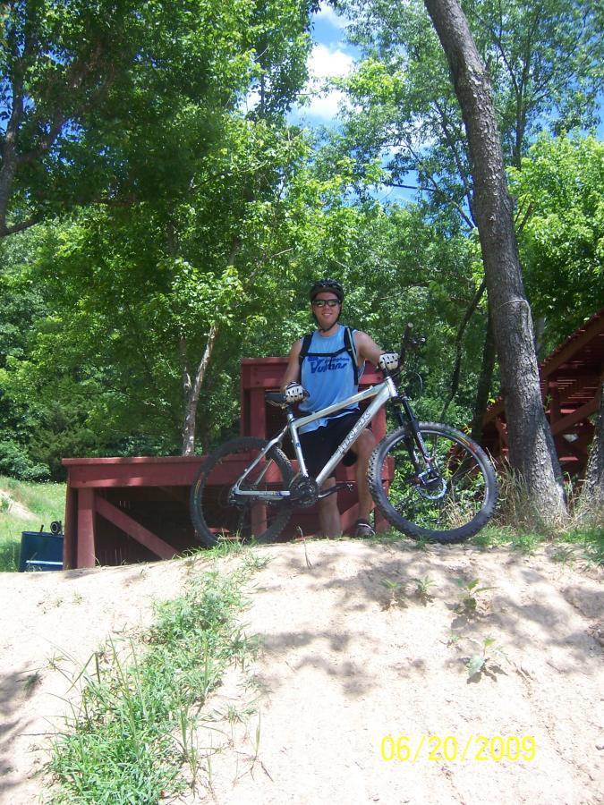 A person standing next to a mountain bike on a dirt hill, with lush green trees in the background. The individual is wearing a blue tank top, a helmet, and gloves, and appears to be enjoying a sunny day outdoors. In the background, there is a wooden structure partially visible.  Slaughter Pen Trail mountain bike trail.