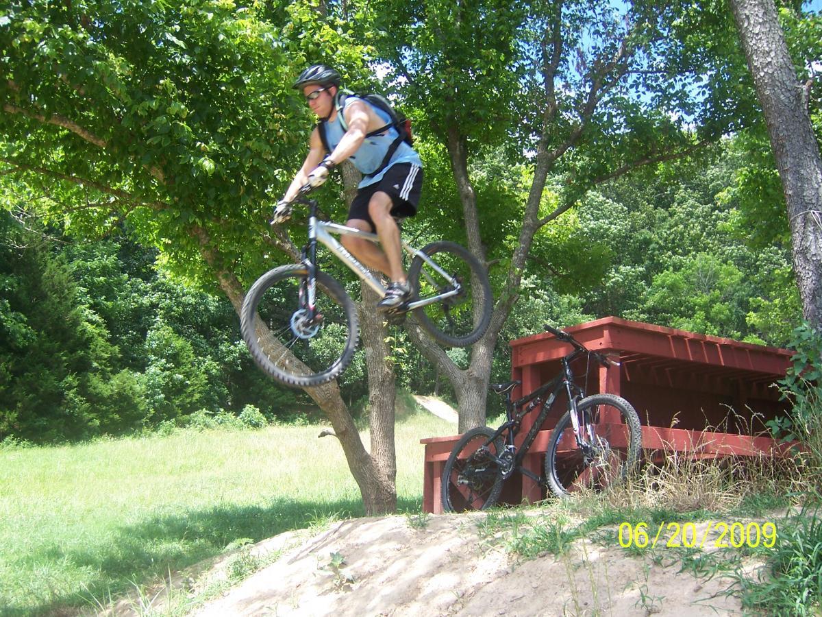 A mountain biker mid-air, performing a jump off a dirt ramp with trees and grass in the background. A second mountain bike is resting on a red wooden structure nearby. Slaughter Pen Trail mountain bike trail.