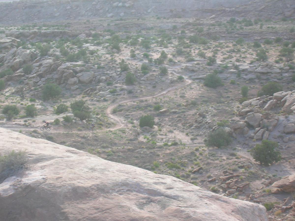A scenic view of a rocky landscape with a winding dirt path. A mountain biker is seen riding along the trail in the background, surrounded by sparse vegetation and rocky outcrops under a hazy sky. Sovereign Single Track mountain bike trail.