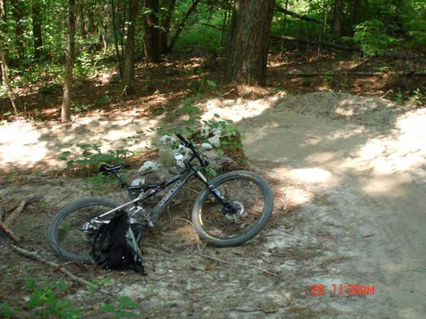 A mountain bike lying on the ground near a dirt path in a wooded area, surrounded by trees and underbrush. A black backpack is positioned next to the bike, and a small dirt jump can be seen in the background. Forks Area Trail System (FATS) mountain bike trail.