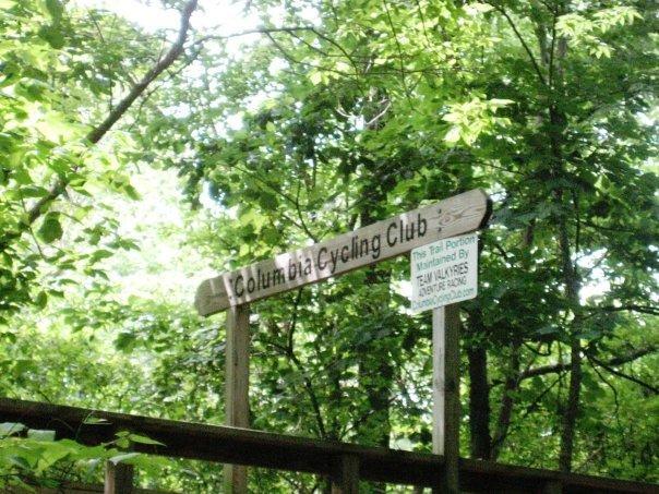 A wooden sign reading "Columbia Cycling Club" surrounded by lush green trees and foliage, with an additional sign below indicating maintenance information. Chickasaw Trace mountain bike trail.