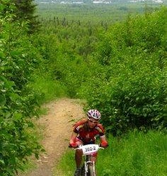 A young cyclist wearing a helmet and a red and black jersey rides a mountain bike along a rocky trail surrounded by green foliage, with a distant city skyline visible in the background. Hillside / Far North Bicentennial Park mountain bike trail.