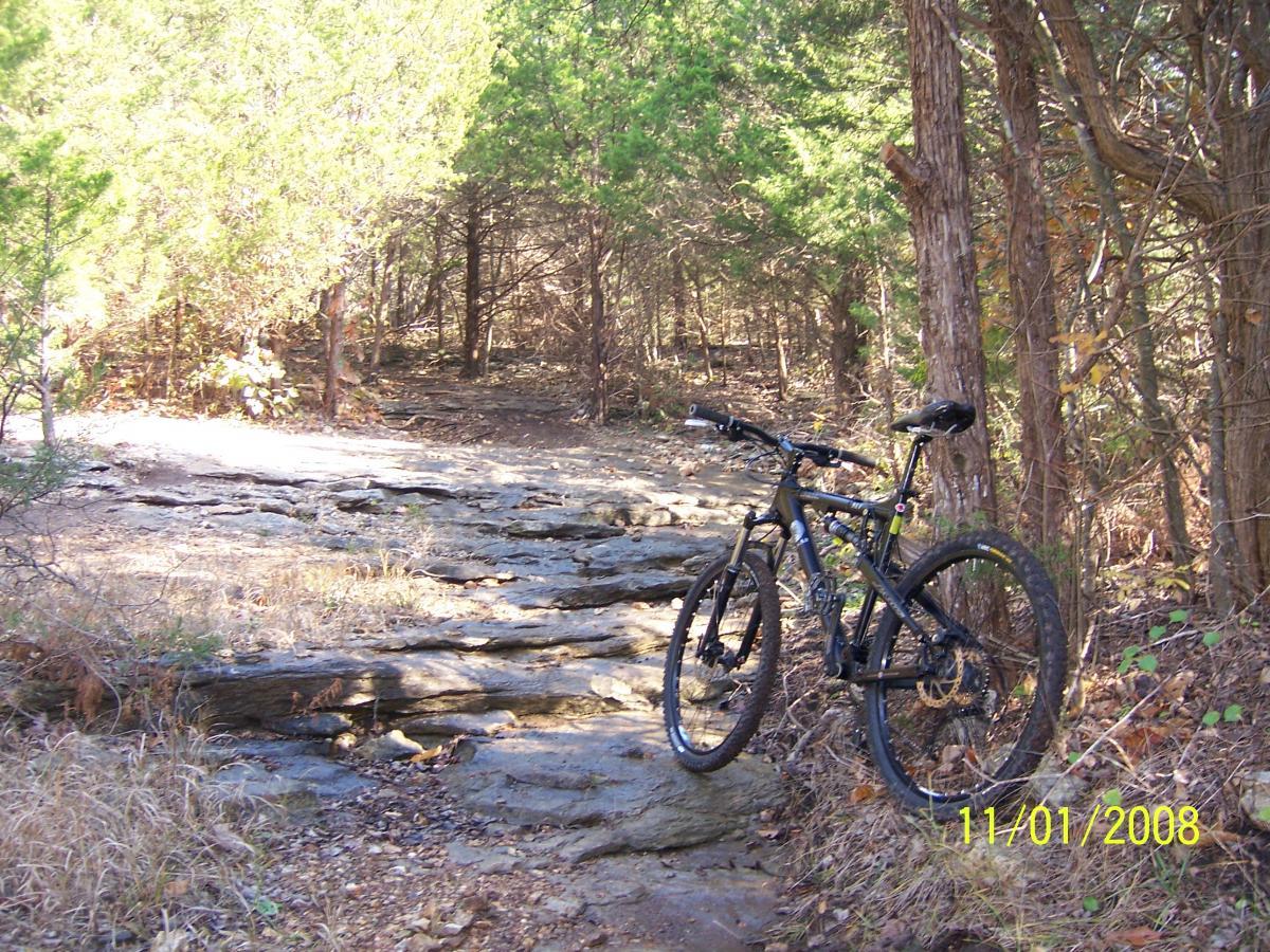A mountain bike leaning against a rocky trail surrounded by greenery and trees. The path is rugged and natural, indicating a scenic outdoor location suitable for biking. Sac River Trail mountain bike trail.