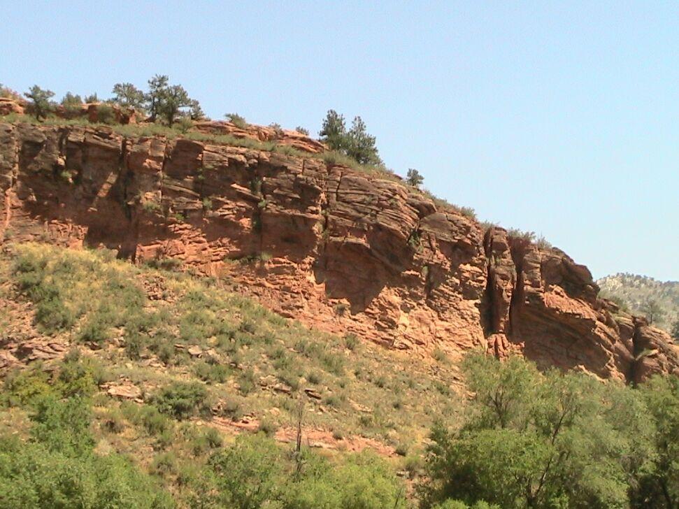 A rocky cliffside featuring reddish-brown stone formations and patches of green vegetation, under a clear blue sky. Bobcat Ridge mountain bike trail.
