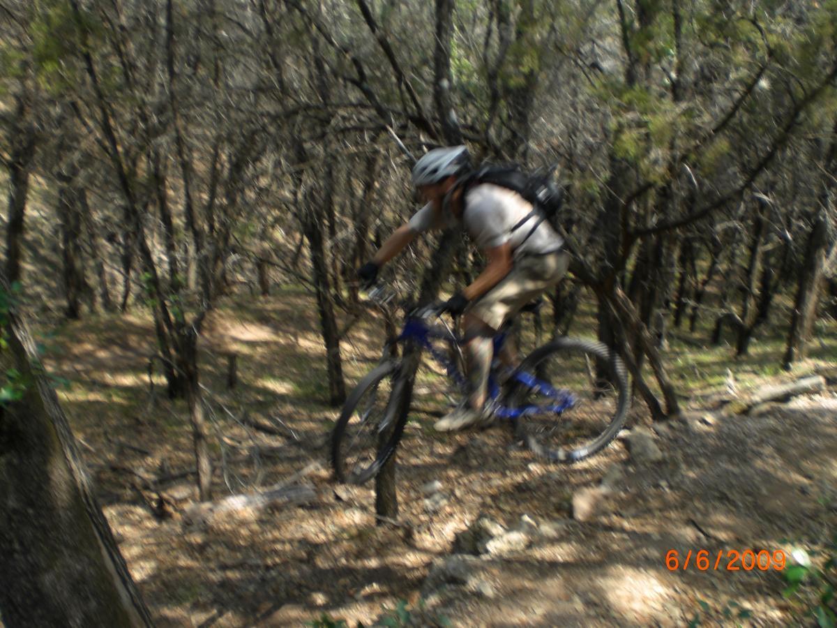 A mountain biker in mid-air navigating a rugged trail surrounded by trees, demonstrating skilled descent along a forest path. The motion is captured with a slight blur, conveying the speed and action of the ride. The date is displayed in the corner, indicating the photo was taken on June 6, 2009. Muleshoe Bend LCRA mountain bike trail.