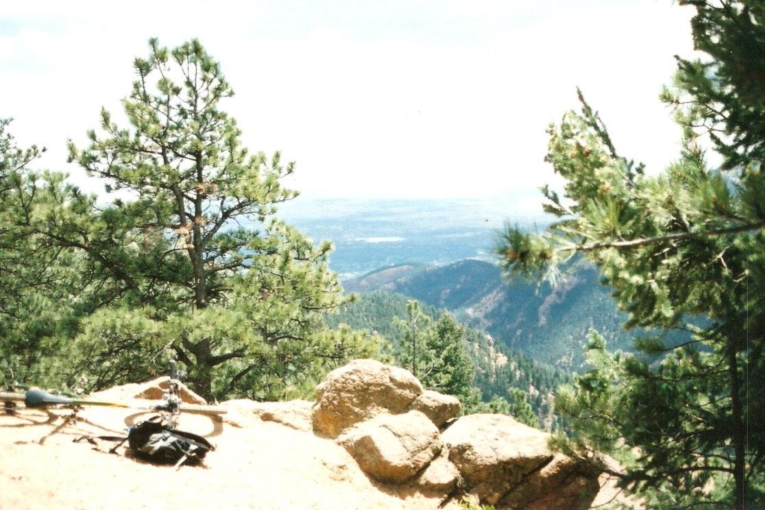 A scenic view from a mountain viewpoint, featuring lush green pine trees and rocky outcrops, with a distant valley and hills visible in the background under a bright sky. A hiking backpack and some equipment are positioned on the ground in the foreground. Captain Jack's mountain bike trail.
