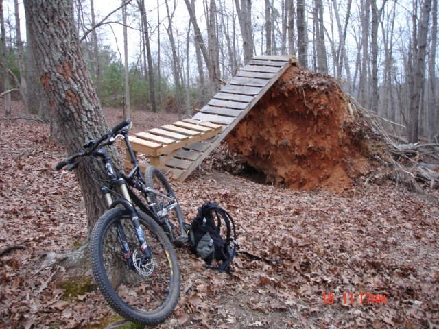A mountain bike leaning against a tree in a wooded area, next to a wooden ramp leading up to an elevated platform made of planks. In the background, there is an uprooted tree and a carpet of fallen leaves on the ground. Georgia International Horse Park mountain bike trail.