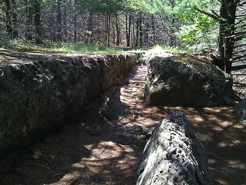 A narrow, natural pathway runs between two large, rocky formations in a forested area, with trees and greenery visible in the background. Sunlight filters through the branches, casting dappled light on the ground. Fancy Creek State Park mountain bike trail.