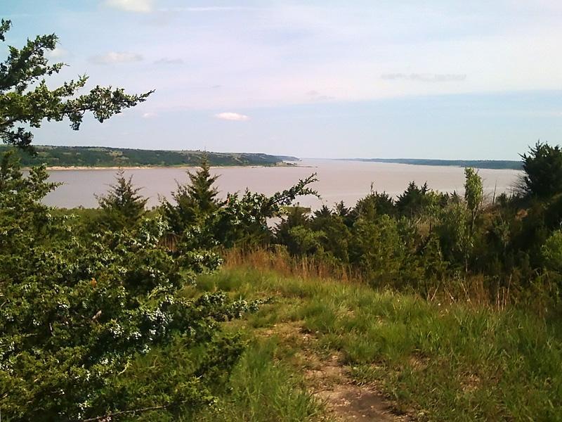 A scenic view of a river surrounded by green vegetation, with rolling hills visible in the background under a partly cloudy sky. The foreground features a variety of shrubs and grasses. Fancy Creek State Park mountain bike trail.