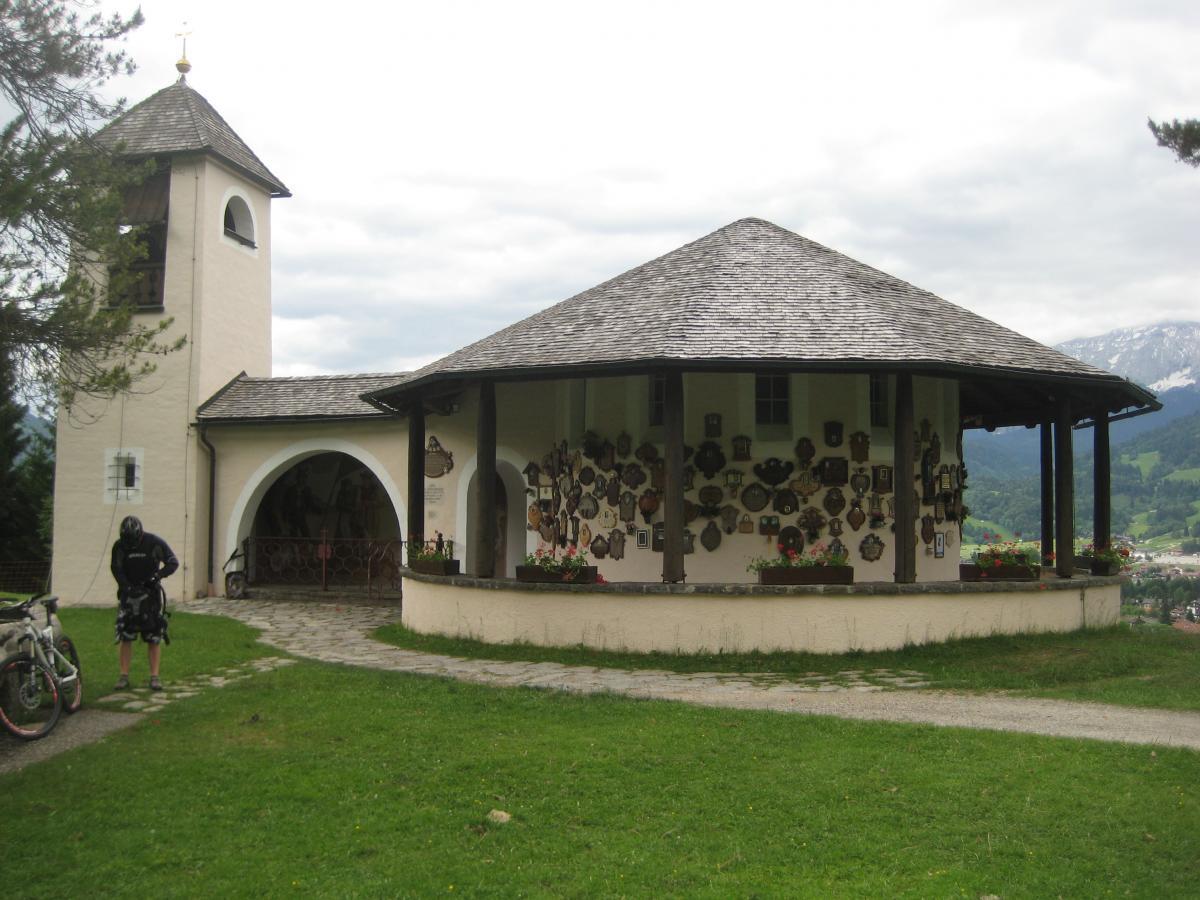 A small church with a round structure and a pointed tower, surrounded by a grassy area, featuring an array of decorative items on its exterior walls. A person in cycling gear stands nearby, with a bicycle parked beside them. The background includes rolling hills and a cloudy sky. Kramer Plateauweg mountain bike trail.