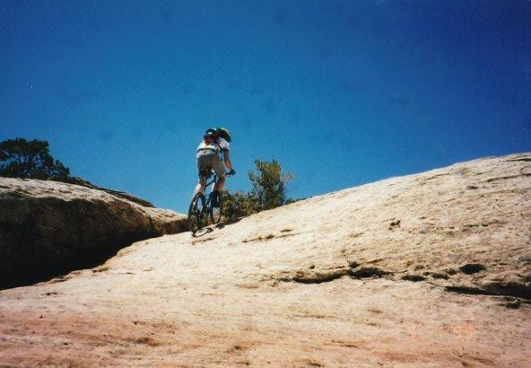 A person riding a mountain bike uphill on a rocky terrain under a clear blue sky. The scene captures the adventurous spirit of outdoor biking amidst natural surroundings. Gooseberry Mesa mountain bike trail.