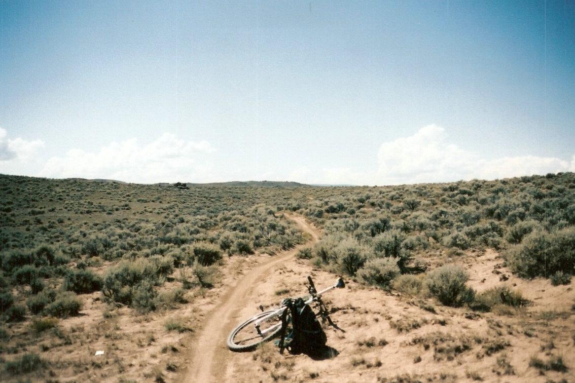 A mountain bike lying on the dirt path in a sparse, grassy landscape under a clear blue sky, surrounded by low shrubs and rolling hills. Hartman Rocks mountain bike trail.