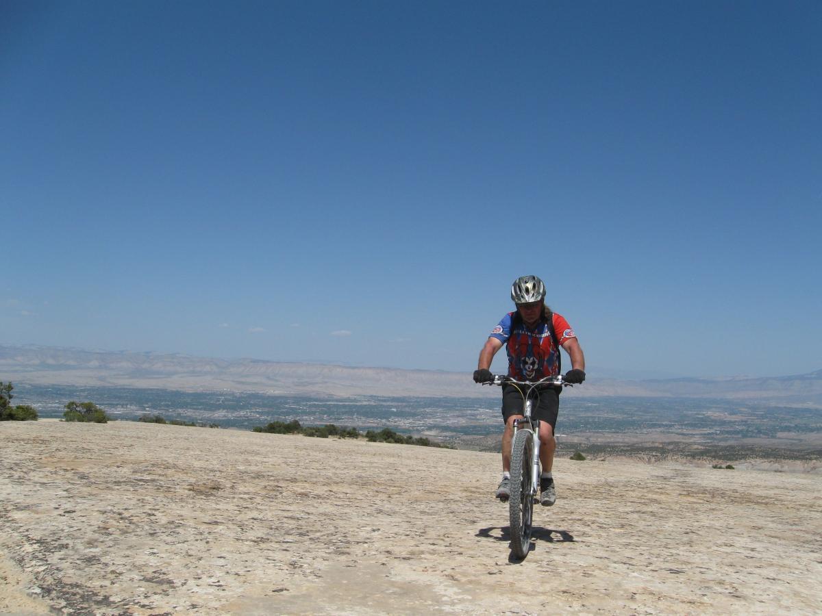 A person wearing a helmet and a colorful cycling jersey is mountain biking across a rocky terrain with a panoramic view of distant mountains and valleys under a clear blue sky. The Ribbon mountain bike trail.