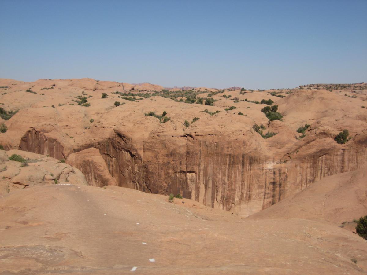 A panoramic view of a sandy desert landscape featuring gently rolling hills and rock formations under a clear blue sky. Sparse vegetation, including small shrubs, can be seen intermittently across the terrain. Slickrock mountain bike trail.