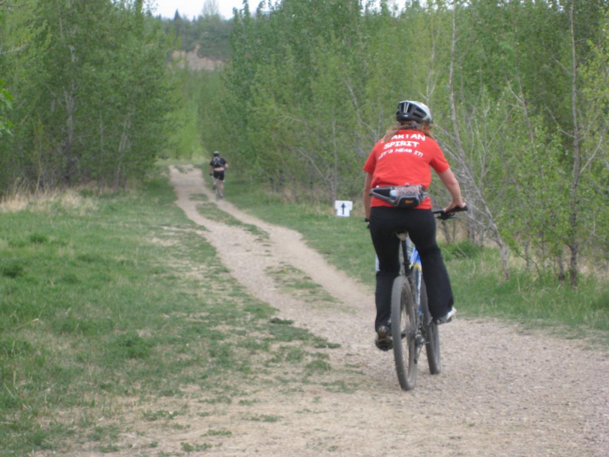 A person riding a mountain bike on a gravel path surrounded by greenery, with another cyclist in the background. The rider in front is wearing a red shirt and a black helmet. Terwillegar Park mountain bike trail.