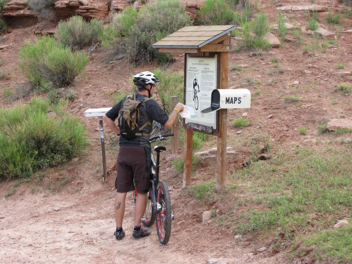 A mountain biker examines a trail map at an information board in a natural setting, surrounded by shrubs and reddish soil. The board provides safety information and details about the trails, while a mailbox labeled "MAPS" is nearby. Porcupine Rim mountain bike trail.