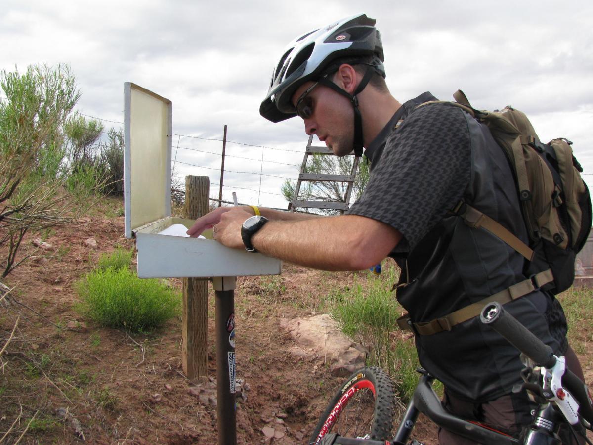 A cyclist in a helmet and sunglasses is leaning over a small metal box mounted on a post, looking inside. He is wearing a black shirt and a backpack, positioned in a rural area with shrubs and a fence in the background under an overcast sky. Porcupine Rim mountain bike trail.