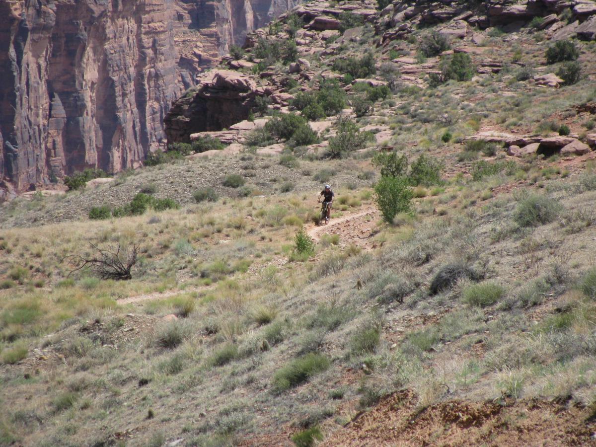 A mountain biker rides along a dirt trail in a rocky, mountainous landscape, surrounded by sparse vegetation and steep cliffs in the background. Porcupine Rim mountain bike trail.