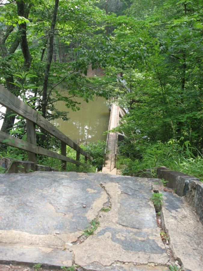A view from the top of a stone staircase leading down to a wooden bridge over a tranquil body of water, surrounded by lush green foliage and trees. Lake Russell Loop mountain bike trail.