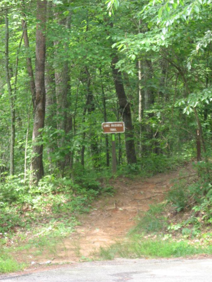A dirt path leading into a lush, green forest, marked by a sign that reads "Lake Russell Trail." The area is surrounded by tall trees and abundant vegetation, creating a serene and inviting atmosphere for outdoor exploration. Lake Russell Loop mountain bike trail.