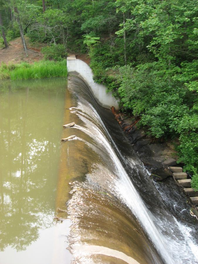 A serene landscape featuring a concrete dam with water cascading over its edge into a calm, muddy river below. Surrounding the dam, lush green trees and vegetation create a natural backdrop. The image captures the flow of water as it spills over the dam into the river, with visible steps leading down the side. Lake Russell Loop mountain bike trail.
